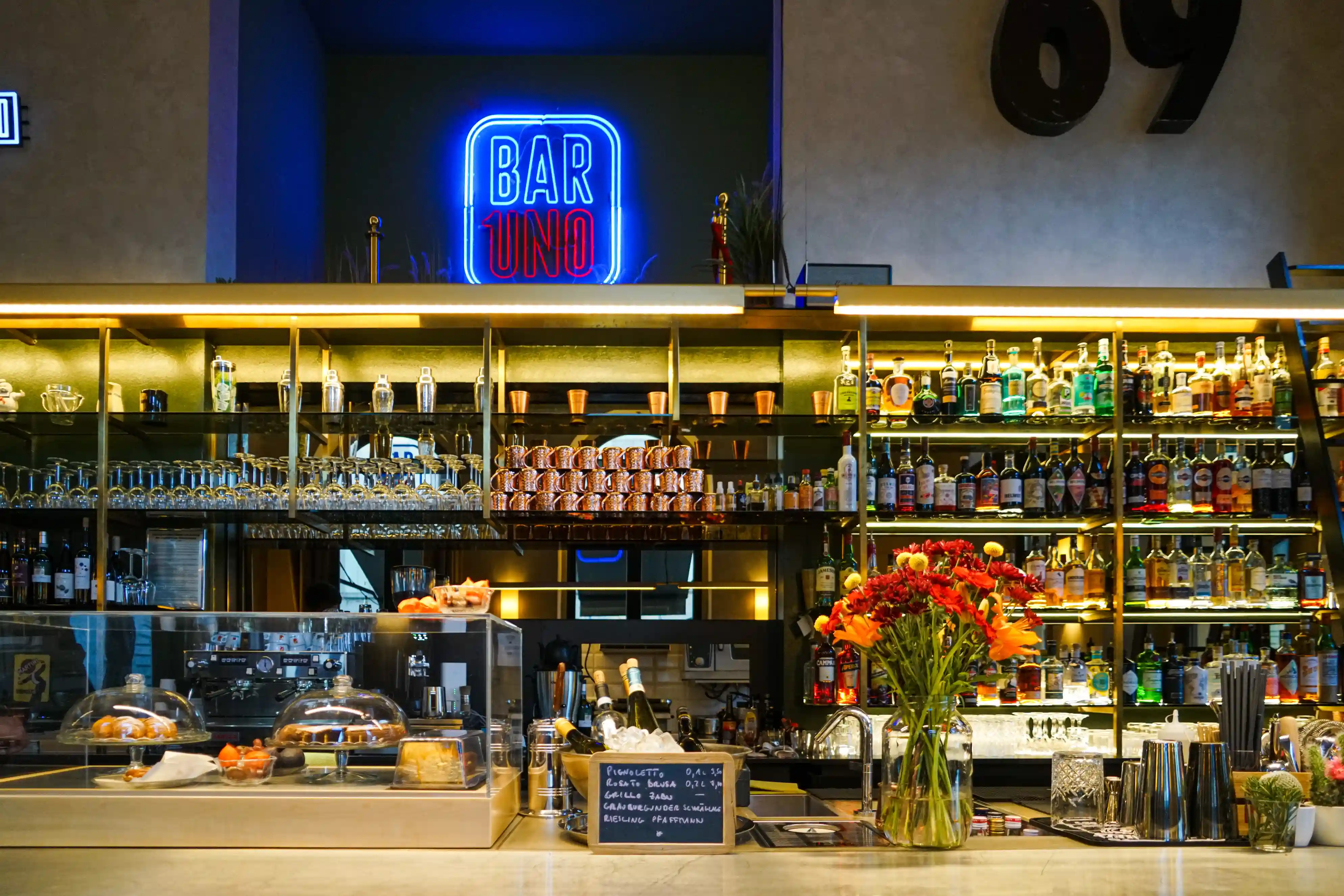 Der Bartresen mit Blumen und Flaschen, vor einer Schrankwand mit Gläsern und Cocktailzutaten